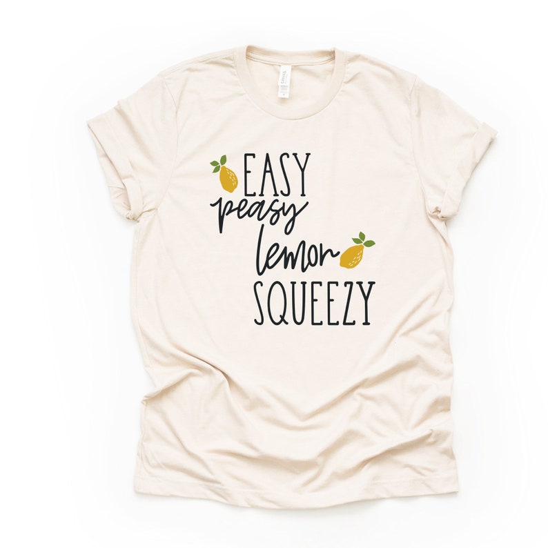 Puede incluir: Una camiseta beige con el texto "EASY peasy lemon SQUEEZY" en letra cursiva negra. Dos limones amarillos con hojas verdes est&aacute;n incluidos en el dise&ntilde;o.