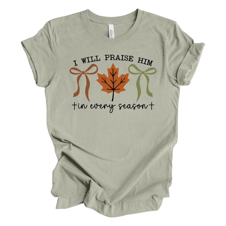May include: A light green t-shirt with the text "I WILL PRAISE HIM in every season+". The design includes a brown bow, an orange maple leaf, and a green bow. The shirt is made of a soft material.