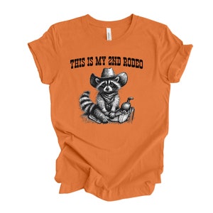 Pode incluir: Camiseta laranja com uma ilustra&ccedil;&atilde;o em preto e branco de um guaxinim usando um chap&eacute;u de caub&oacute;i e sentado em uma sela. O texto "THIS IS MY 2ND RODEO" est&aacute; impresso na camiseta.