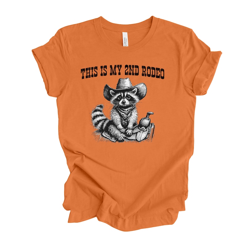 Pode incluir: Camiseta laranja com uma ilustra&ccedil;&atilde;o em preto e branco de um guaxinim usando um chap&eacute;u de caub&oacute;i e sentado em uma sela. O texto "THIS IS MY 2ND RODEO" est&aacute; impresso na camiseta.