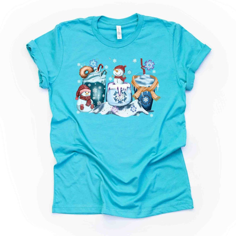 May include: A turquoise blue t-shirt with a winter-themed design featuring three snowmen holding mugs of hot chocolate and a donut. The snowmen are wearing scarves and hats, and the mugs are decorated with snowflakes.