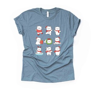 Pode incluir: Uma camiseta azul claro com um gr&aacute;fico de nove ursos polares brancos vestidos com trajes de Natal vermelhos e brancos. Os ursos polares est&atilde;o realizando v&aacute;rias atividades, como segurar um presente, usar uma coroa e patinar no gelo.