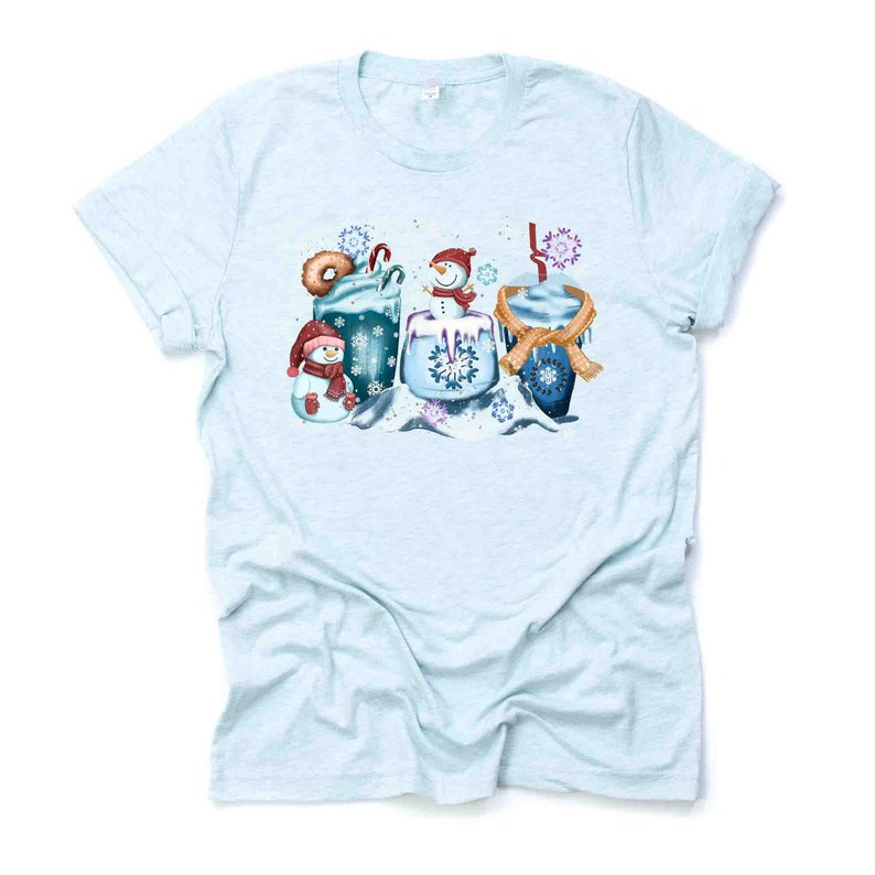 May include: A light blue t-shirt with a winter-themed design featuring three snowmen holding mugs of hot chocolate with whipped cream and candy canes. The design is surrounded by snowflakes.