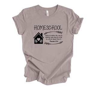 Puede incluir: Una camiseta gris claro con un gr&aacute;fico negro que dice "HOMESCHOOL" con una casa, flores y una flecha. El texto debajo del gr&aacute;fico dice "Instruye al ni&ntilde;o en el camino en que debe andar, y aun cuando fuere viejo no se apartar&aacute; de &eacute;l. Proverbios 22:6"