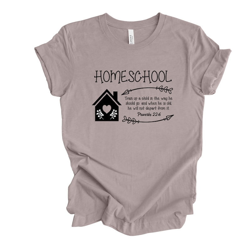 Puede incluir: Una camiseta gris claro con un gr&aacute;fico negro que dice "HOMESCHOOL" con una casa, flores y una flecha. El texto debajo del gr&aacute;fico dice "Instruye al ni&ntilde;o en el camino en que debe andar, y aun cuando fuere viejo no se apartar&aacute; de &eacute;l. Proverbios 22:6"