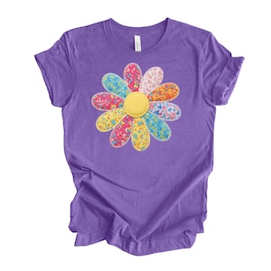 May include: A purple t-shirt featuring a large, colorful daisy design. The daisy has petals in various colors, including pink, blue, yellow, and orange, with floral patterns. The center of the daisy is a solid yellow circle. The shirt is made of a soft material.