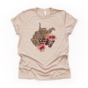 May include: A beige t-shirt with a leopard print outline of the state of West Virginia with floral accents and the letters "WV" in a colorful striped pattern.