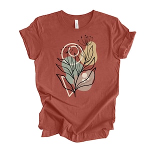 May include: A rust-colored t-shirt with a black and white abstract floral design. The design features a stylized flower with leaves and geometric shapes in shades of beige, green, and red.