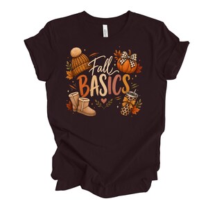 May include: A dark brown short-sleeve t-shirt featuring a graphic print with the text Fall BASICS. The design includes a tan knit beanie, an orange pumpkin with a buffalo plaid bow, tan boots, and a leopard print tumbler, surrounded by autumn leaves.