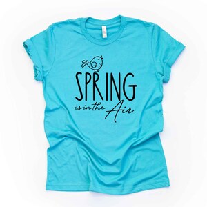 May include: A turquoise blue t-shirt with a black graphic of a bird and the text "Spring is in the Air".