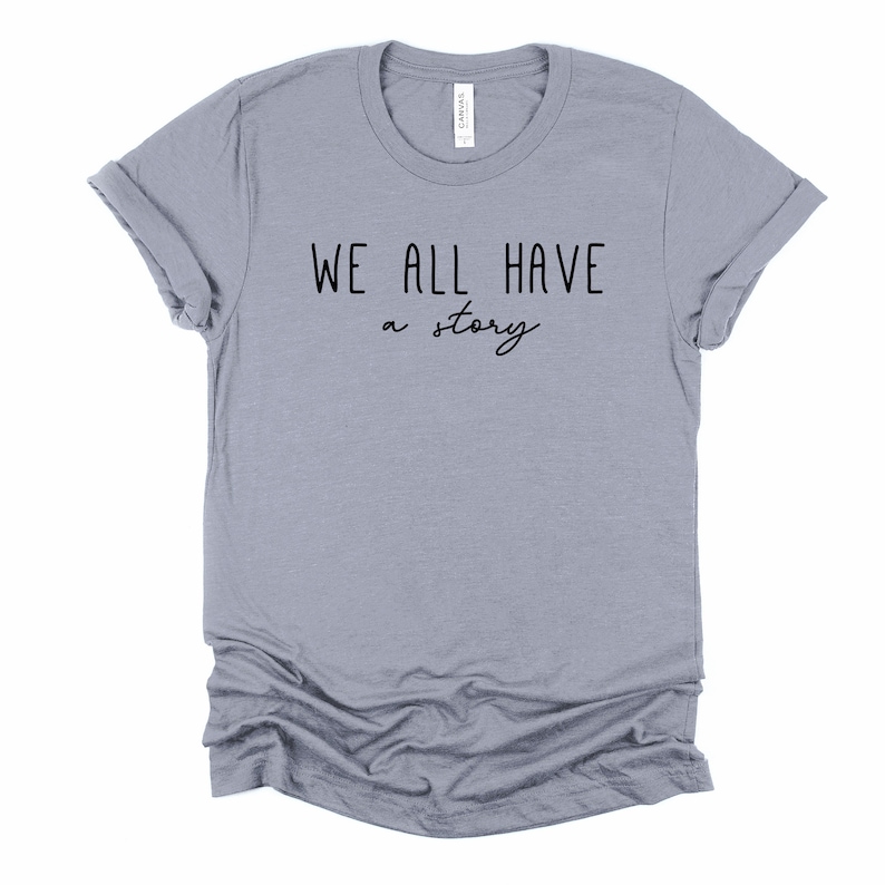 May include: A heather grey t-shirt with the text "WE ALL HAVE a story" printed in black.