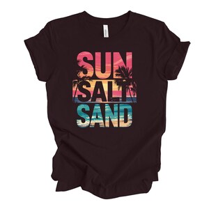 Op de afbeelding: Een donkerbruin t-shirt met de woorden "SUN SALT SAND" in een kleurrijk, gelaagd ontwerp. De tekst bevat een zonsondergang en palmbomen, die een strandthema oproepen. Het shirt is gemaakt van een zacht materiaal.