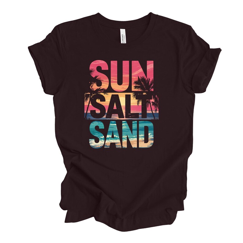 Op de afbeelding: Een donkerbruin t-shirt met de woorden "SUN SALT SAND" in een kleurrijk, gelaagd ontwerp. De tekst bevat een zonsondergang en palmbomen, die een strandthema oproepen. Het shirt is gemaakt van een zacht materiaal.