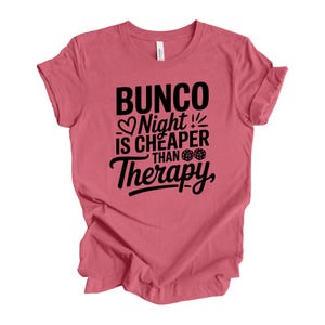 May include: A dusty rose t-shirt with the text "Bunco Night is Cheaper Than Therapy" in black. The design includes a heart, dice, and a stylized font. The shirt is made of a soft material and has short sleeves.