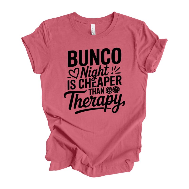K&ouml;nnte beinhalten: Staubrosafarbenes T-Shirt mit dem schwarzen Text "Bunco Night is Cheaper Than Therapy". Das Design beinhaltet ein Herz, W&uuml;rfel und eine stilisierte Schriftart. Das Shirt besteht aus weichem Material und hat kurze &Auml;rmel.
