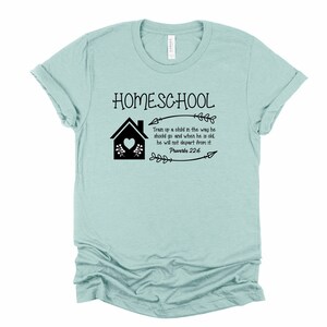 Puede incluir: Una camiseta azul claro con el texto "HOMESCHOOL" en negro. La camiseta tambi&eacute;n presenta una casa negra con un coraz&oacute;n, flores y flechas. El texto "Train up a child in the way he should go and when he is old, he will not depart from it. Proverbs 22:6" tambi&eacute;n est&aacute; impreso en negro.