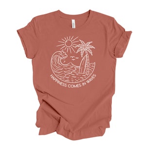 May include: A rust-colored t-shirt with a white graphic design of a sun, palm tree, surfboard, and wave. The text "Happiness comes in waves" is written below the graphic.