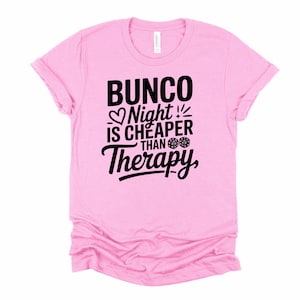 K&ouml;nnte beinhalten: Hellrosa T-Shirt mit dem schwarzen Text "Bunco Night is Cheaper Than Therapy". Das Design beinhaltet ein Herz, W&uuml;rfel und Ausrufezeichen. Das T-Shirt hat einen Rundhalsausschnitt und hochgekrempelte &Auml;rmel.
