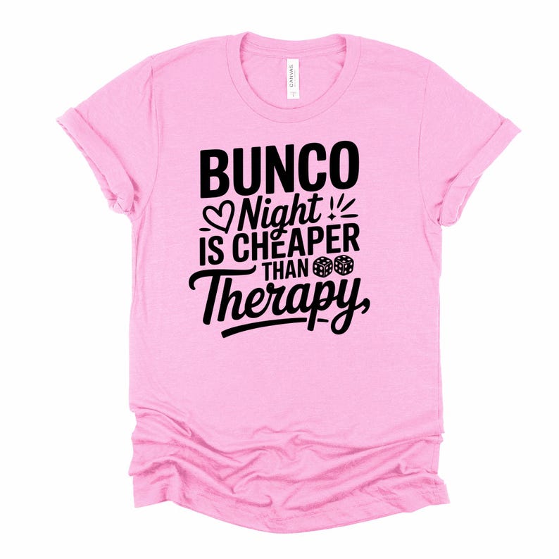K&ouml;nnte beinhalten: Hellrosa T-Shirt mit dem schwarzen Text "Bunco Night is Cheaper Than Therapy". Das Design beinhaltet ein Herz, W&uuml;rfel und Ausrufezeichen. Das T-Shirt hat einen Rundhalsausschnitt und hochgekrempelte &Auml;rmel.