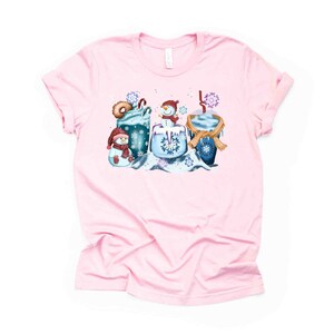 May include: Pink t-shirt with a winter-themed design featuring a snowman, a mug of hot chocolate, and a donut. The design is in shades of blue, brown, and white.