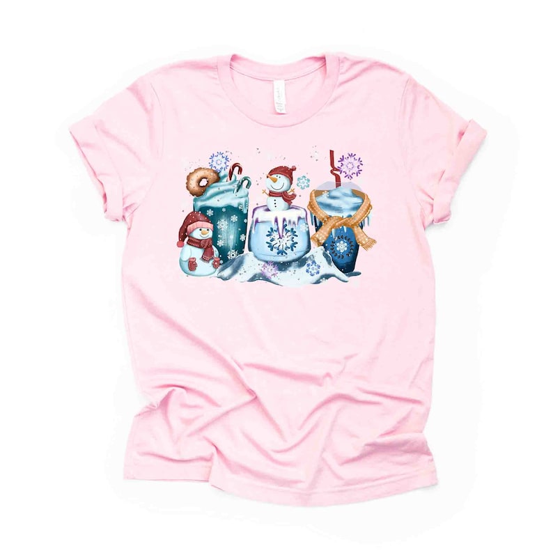 May include: Pink t-shirt with a winter-themed design featuring a snowman, a mug of hot chocolate, and a donut. The design is in shades of blue, brown, and white.