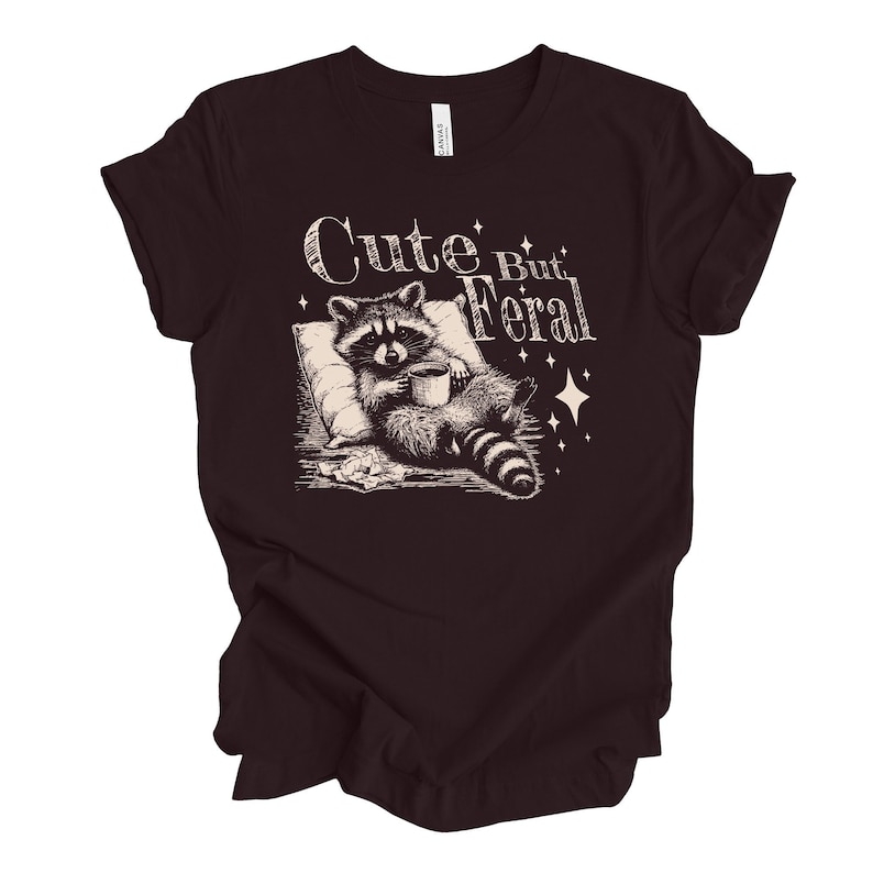 May include: A dark brown t-shirt with a graphic of a raccoon holding a cup of coffee and the text "Cute But Feral" in a vintage style font.