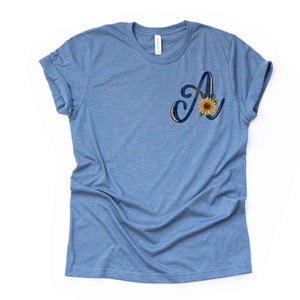 May include: A blue t-shirt with a sunflower and the letter "A" in a navy blue script font.