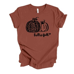 May include: A rust-colored t-shirt featuring a black graphic of two pumpkins. One pumpkin has a leopard print design, and the other is a solid outline. The words "hello fall" are written below the pumpkins in a script font, with a heart symbol.