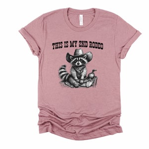 Pode incluir: Uma camiseta rosa com uma ilustra&ccedil;&atilde;o em preto e branco de um guaxinim usando um chap&eacute;u de cowboy e montando uma sela. O texto "THIS IS MY 2ND RODEO" est&aacute; impresso na camiseta.