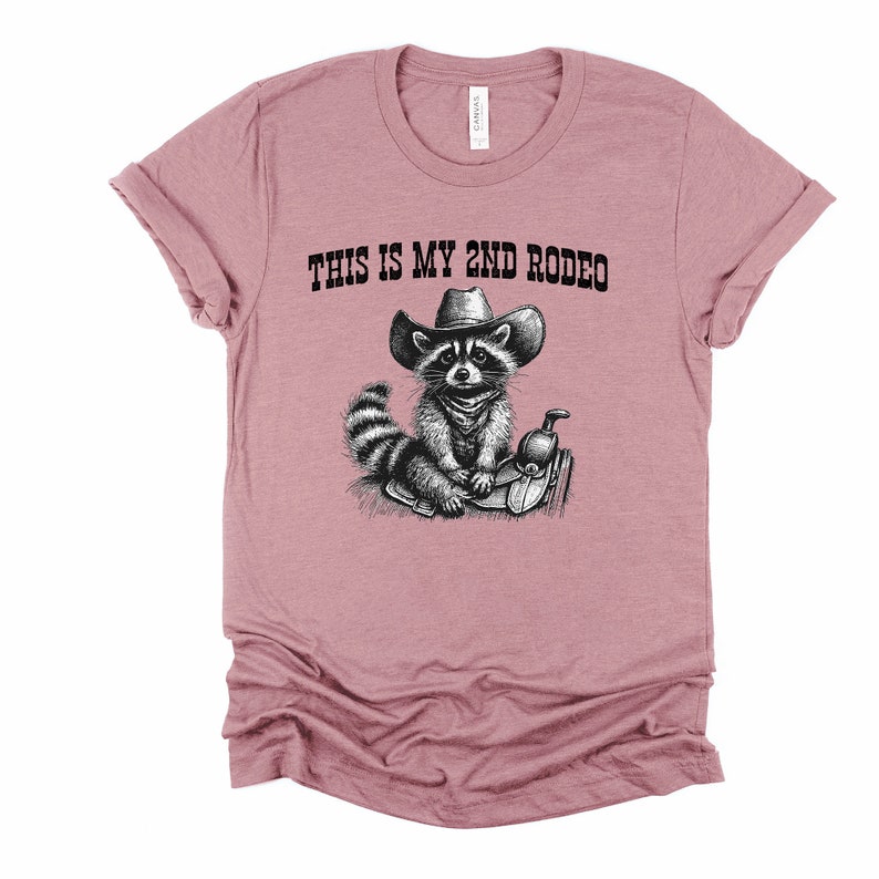 Pode incluir: Uma camiseta rosa com uma ilustra&ccedil;&atilde;o em preto e branco de um guaxinim usando um chap&eacute;u de cowboy e montando uma sela. O texto "THIS IS MY 2ND RODEO" est&aacute; impresso na camiseta.