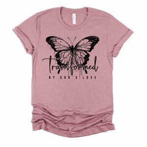 Peut inclure: Un t-shirt mauve avec un motif de papillon noir et le texte "transform&eacute; par l'amour de Dieu".