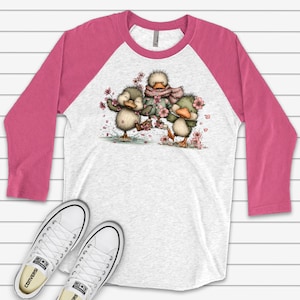 May include: A white and heather gray raglan shirt with pink sleeves. The shirt features a whimsical illustration of three cartoon ducklings surrounded by pink flowers. White Converse sneakers are at the bottom of the image.