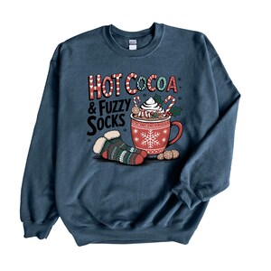 May include: A navy blue sweatshirt with a graphic of a mug of hot cocoa with whipped cream and candy canes, and a pair of fuzzy socks. The text "Hot Cocoa & Fuzzy Socks" is printed on the sweatshirt.