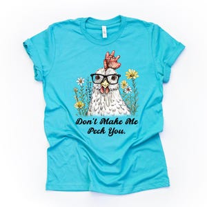 May include: A turquoise t-shirt featuring a whimsical illustration of a chicken wearing glasses, surrounded by flowers. The text "Don't Make Me Peck You" is printed below the chicken. A fun and quirky design.