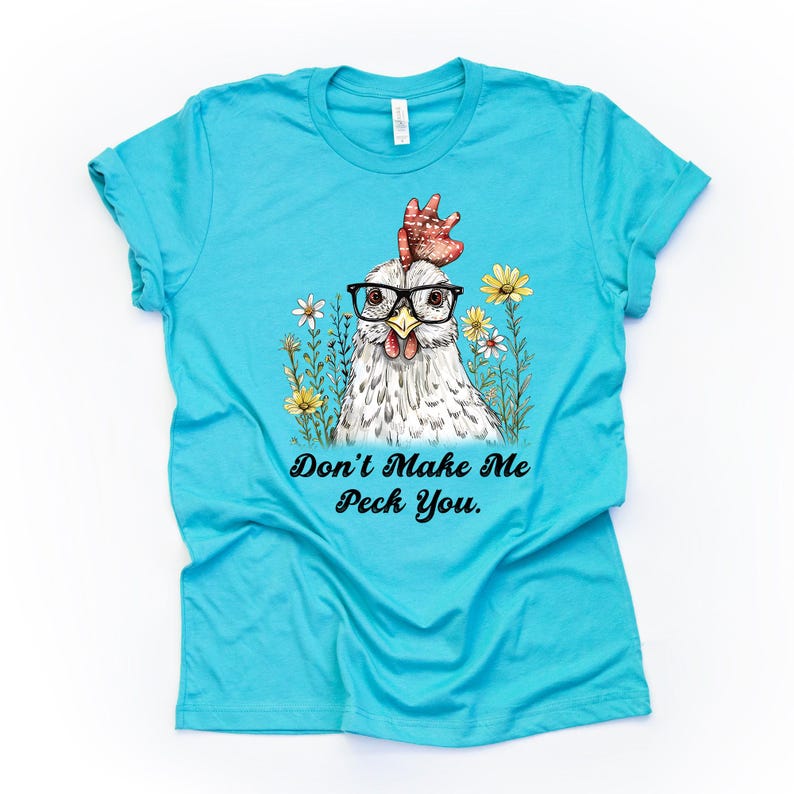 May include: A turquoise t-shirt featuring a whimsical illustration of a chicken wearing glasses, surrounded by flowers. The text "Don't Make Me Peck You" is printed below the chicken. A fun and quirky design.