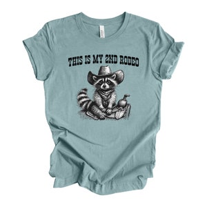 Pode incluir: Uma camiseta azul claro com uma ilustra&ccedil;&atilde;o em preto e branco de um guaxinim usando um chap&eacute;u de cowboy e montando uma sela. O texto "This is my 2nd Rodeo" est&aacute; impresso na camiseta.
