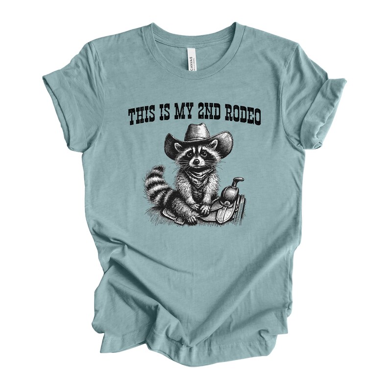 Pode incluir: Uma camiseta azul claro com uma ilustra&ccedil;&atilde;o em preto e branco de um guaxinim usando um chap&eacute;u de cowboy e montando uma sela. O texto "This is my 2nd Rodeo" est&aacute; impresso na camiseta.