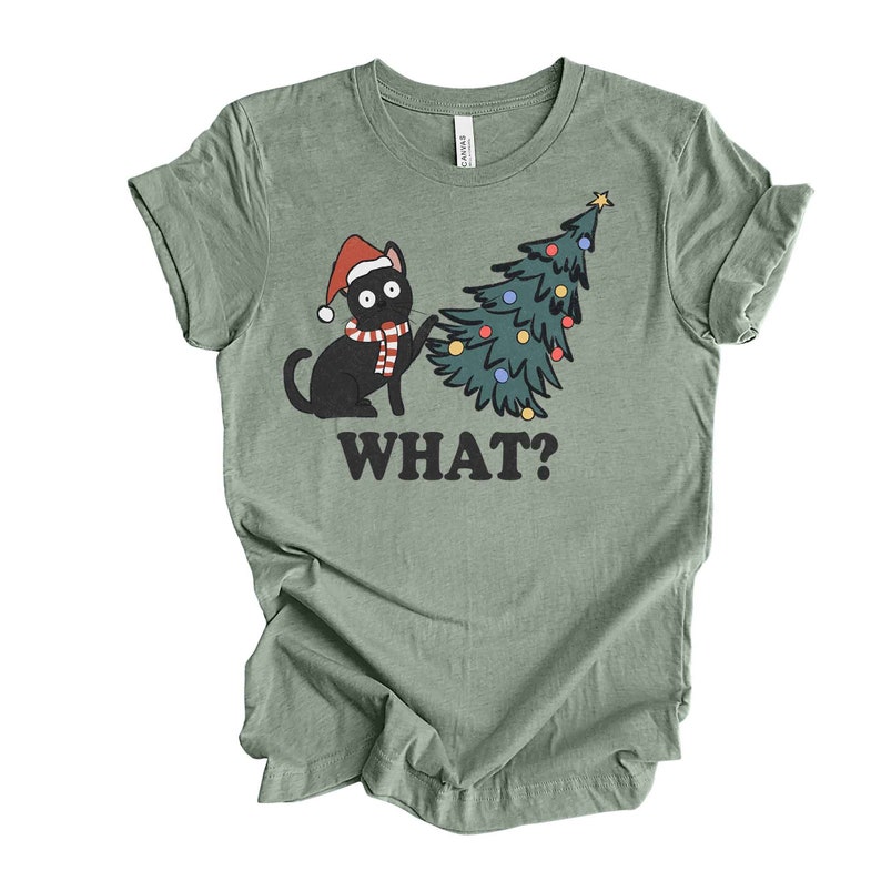 May include: A green t-shirt with a black cat wearing a Santa hat and scarf, looking at a decorated Christmas tree. The text "WHAT?" is printed below the image.