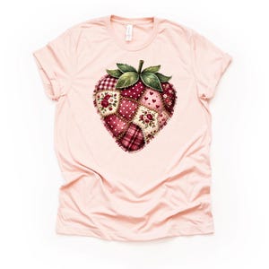 May include: A light pink t-shirt featuring a large, heart-shaped strawberry design. The strawberry is composed of patchwork squares in shades of red, pink, and cream, with floral and polka dot patterns. Green leaves and a stem complete the design.