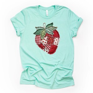 May include: A light turquoise t-shirt featuring a large, appliqued strawberry design. The strawberry is composed of various red fabric patches with white polka dots and floral patterns. The leaves are green and the stem is a twisted brown cord.