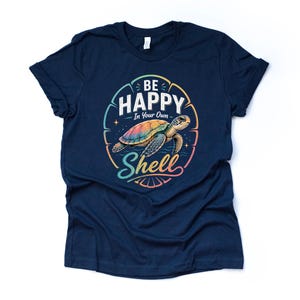 May include: Navy blue t-shirt with a colorful graphic of a sea turtle inside a circular design. The design includes the words "Be Happy In Your Own Shell" in white and a rainbow color scheme. The shirt is made of soft material.