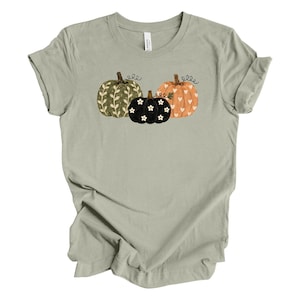May include: A sage green short-sleeve t-shirt featuring a graphic of three pumpkins. The left pumpkin is olive green with white leaf patterns, the center is black with white flower accents, and the right is orange with white heart patterns.