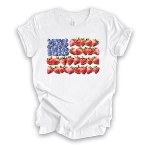 May include: White t-shirt featuring a design of the American flag made of blueberries and strawberries. The flag design is centered on the front of the shirt. The shirt is made of a soft, lightweight material.