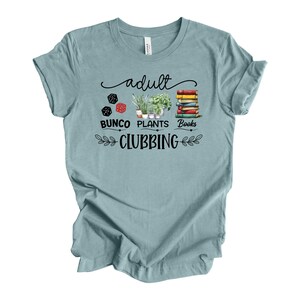 May include: A light blue t-shirt with the words "adult BUNCO PLANTS BOOKS CLUBBING" in black script and a decorative design. The design includes dice, potted plants, and a stack of books.
