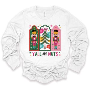 May include: White long-sleeve shirt with a colorful graphic. The graphic features two nutcrackers and a Christmas tree, framed in decorative borders. The text "Y'all are Nuts" is displayed below the images, with star accents.
