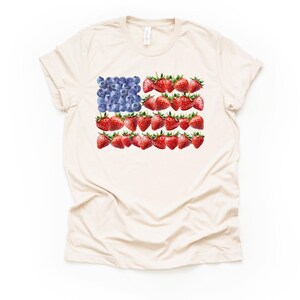 May include: Cream-colored t-shirt featuring a watercolor design of the American flag. The flag's stars are represented by blueberries, and the stripes are made of strawberries. The shirt has short sleeves and a crew neck.