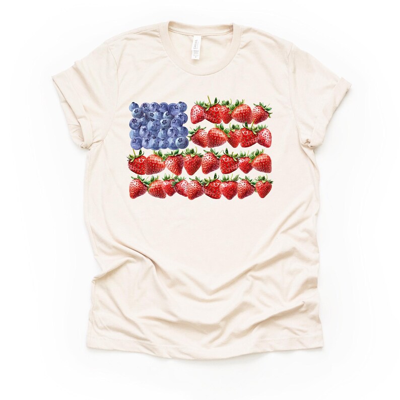 May include: Cream-colored t-shirt featuring a watercolor design of the American flag. The flag's stars are represented by blueberries, and the stripes are made of strawberries. The shirt has short sleeves and a crew neck.