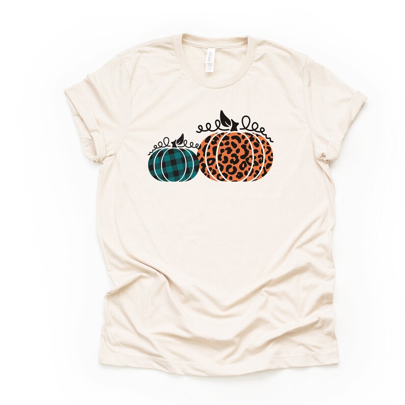 May include: A beige t-shirt with a graphic of two pumpkins. One pumpkin is orange with a black leopard print and the other is teal with a black and white plaid pattern.