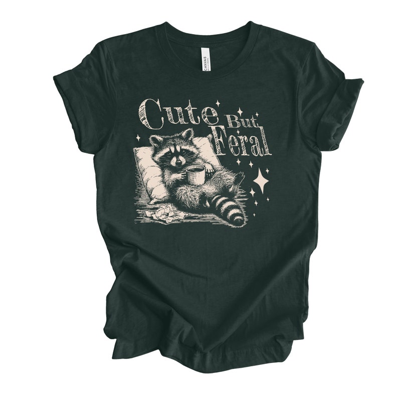 May include: A dark green t-shirt with a graphic of a raccoon holding a cup of coffee and the text "Cute But Feral".