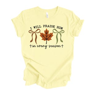 May include: A light yellow t-shirt with the text "I WILL PRAISE HIM in every season" in black. The design includes a brown bow, a brown and orange leaf, and a green bow.